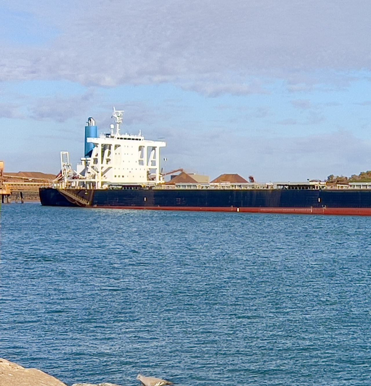 A bulk carrier berthed at a loading terminal in Port Hedland harbour, Western Australia, with ore stockpiles behind it.
